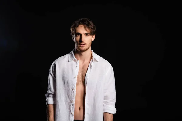 Seductive man in unbuttoned white shirt looking at camera isolated on black — Stock Photo