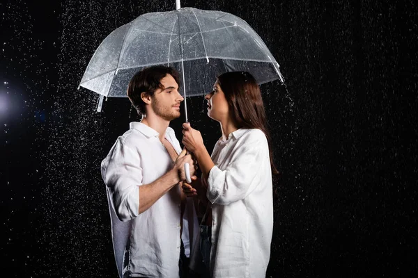 Young adult boyfriend and girlfriend standing in rain with umbrella on black background — Stock Photo