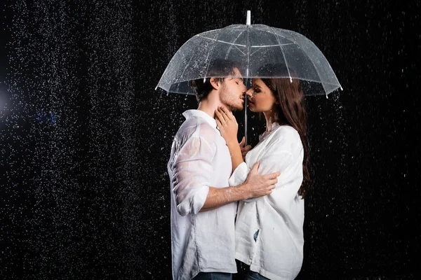 Attractive young adult boyfriend and girlfriend standing in rain with umbrella and kissing on black background — Stock Photo