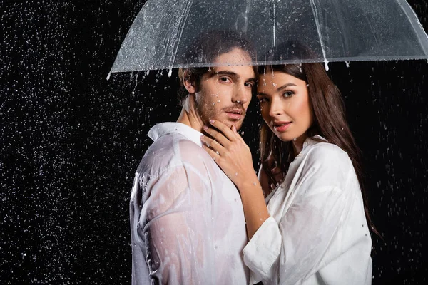 Romantic young adult couple standing in rain with umbrella and looking at camera on black background — Stock Photo
