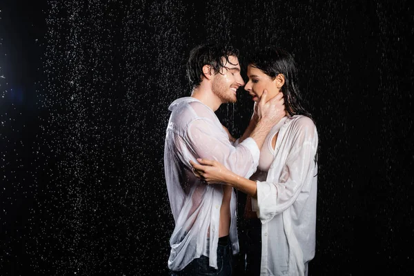 Copain heureux et petite amie étreignant dans des vêtements humides sous la pluie sur fond noir — Photo de stock