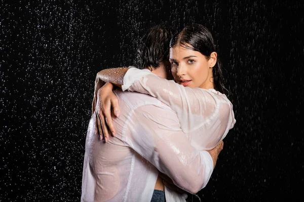 Пристрасна сексуальна пара в мокрому одязі, що стоїть в краплі дощу і обіймається на чорному тлі — стокове фото