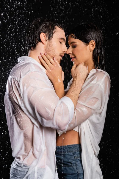 Wet passionate couple holding hands near faces in rain drops on black background — Stock Photo