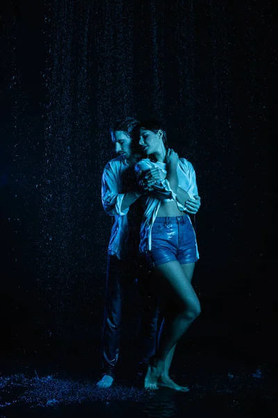 Homem de boa aparência gentilmente abraçando mulher sensual molhada em gotas de água no fundo preto com filtro de cor azul — Fotografia de Stock