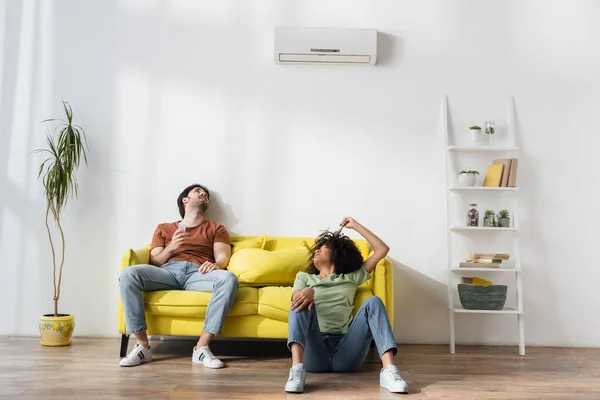 Young interracial couple suffering from heat and looking at air conditioner in modern living room — Stock Photo