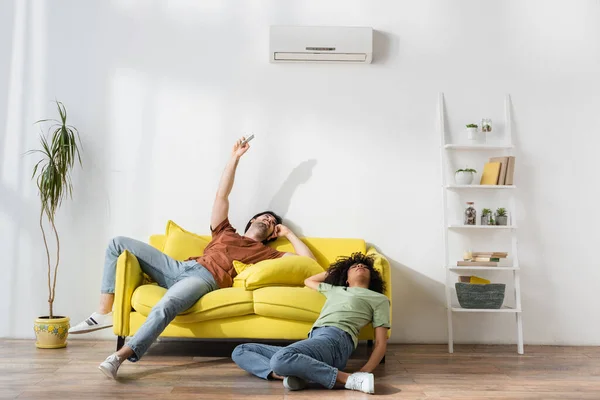 Young man holding remote controller near air conditioner while suffering from heat with african american girlfriend — Stock Photo