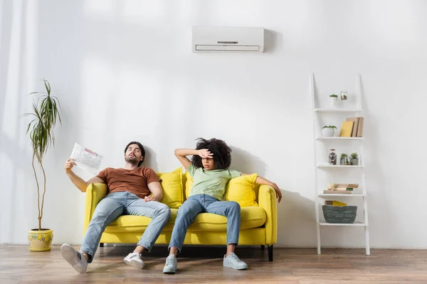 Young man waving newspaper while sitting on couch with african american girlfriend and suffering from heat — Stock Photo