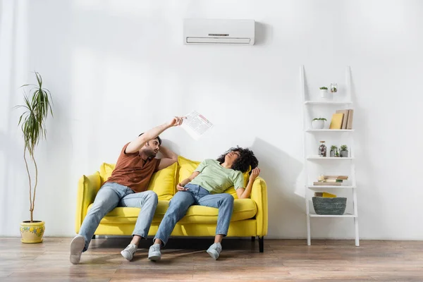 Young man waving newspaper at african american girlfriend while suffering from heat — Stock Photo