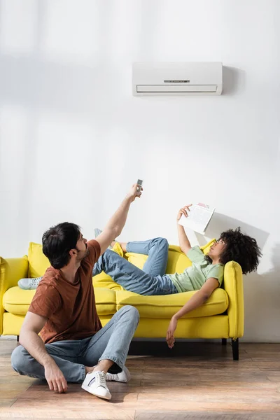 Man holding remote controller near air conditioner and african american girlfriend waving newspaper while suffering from heat — Stock Photo
