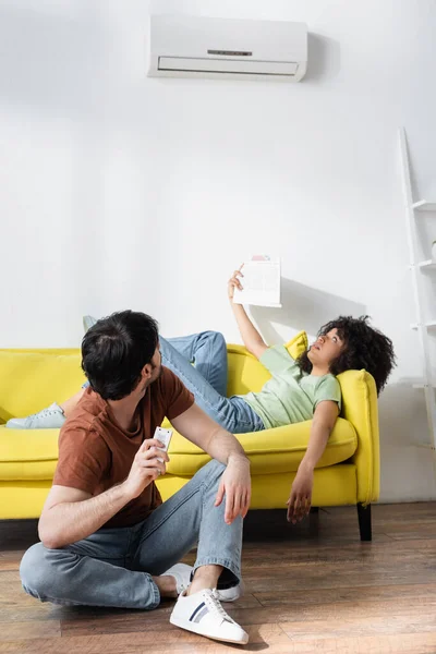 Man holding remote controller and looking at air conditioner near african american girlfriend waving newspaper while suffering from heat — Stock Photo
