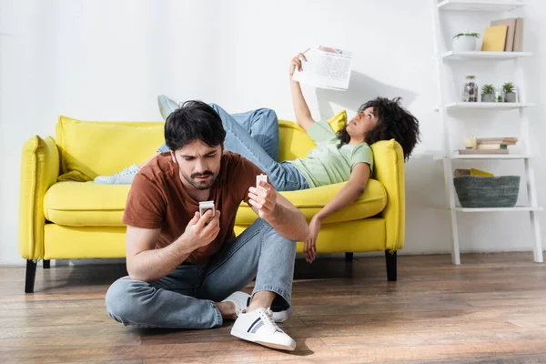 Hombre sosteniendo mando a distancia cerca de afroamericano novia agitando periódico mientras que sufre de calor — Stock Photo