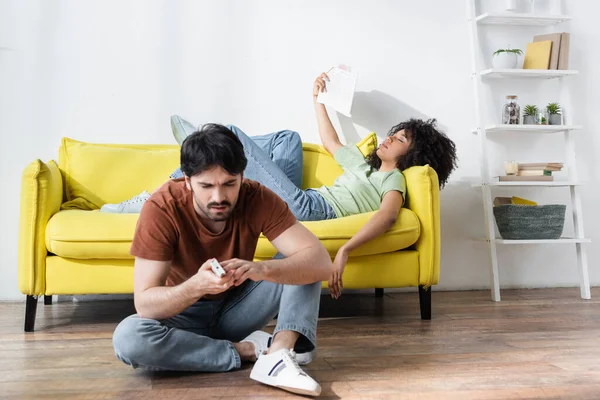 Man fixing remote controller near african american girlfriend waving newspaper while suffering from heat — Stock Photo