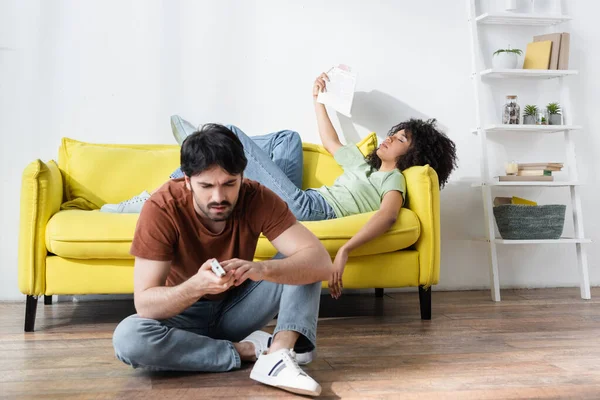 Mann repariert Fernbedienung in der Nähe von afrikanisch-amerikanischer Freundin winkt Zeitung, während sie unter Hitze leidet — Stockfoto