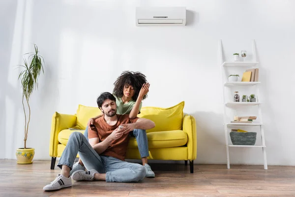 Displeased interracial couple looking at remote controller while sitting on couch near air conditioner — Stock Photo