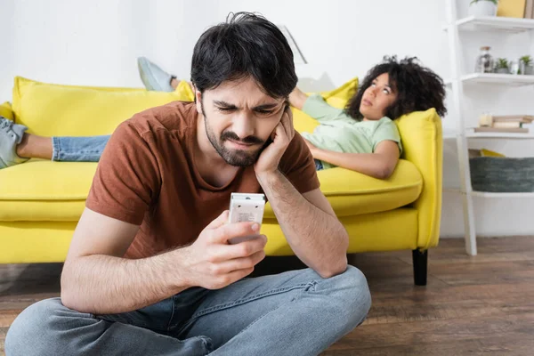 Upset man looking at broken remote controller near blurred african american girlfriend suffering from heat — Stock Photo