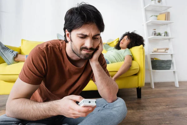 Sad man looking at broken remote controller near blurred african american girlfriend suffering from heat — Stock Photo