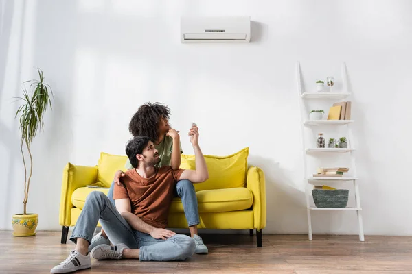 Displeased interracial couple looking at air conditioner while sitting on sofa — Stock Photo