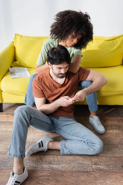 Triste casal interracial olhando para controlador remoto enquanto sentado no sofá perto de ar condicionado — Fotografia de Stock