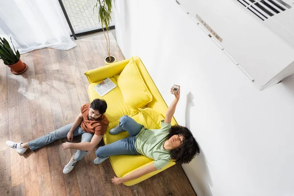 Vista superior de la mujer afroamericana utilizando el controlador remoto cerca del aire acondicionado y novio en el sofá - foto de stock