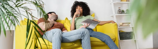 Mann mit Zeitung und Blick auf Afroamerikanerin, die im Wohnzimmer Wasser trinkt, Banner — Stockfoto