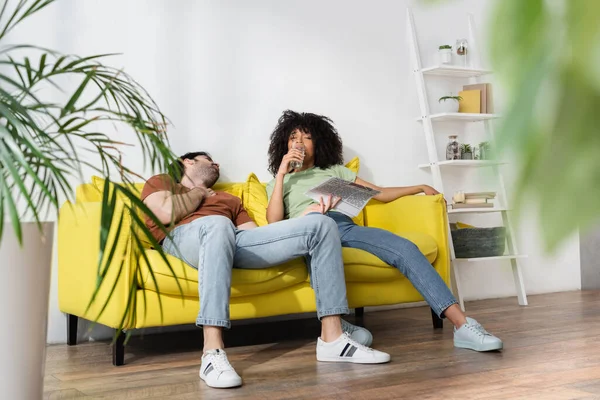 Homem segurando jornal e olhando para a mulher afro-americana bebendo água na sala de estar turva — Fotografia de Stock