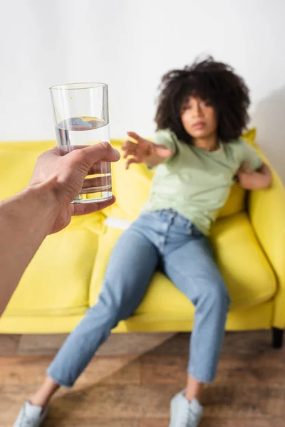 Blurred and curly african american woman with outstretched hand reaching glass of water in male hand — Stock Photo