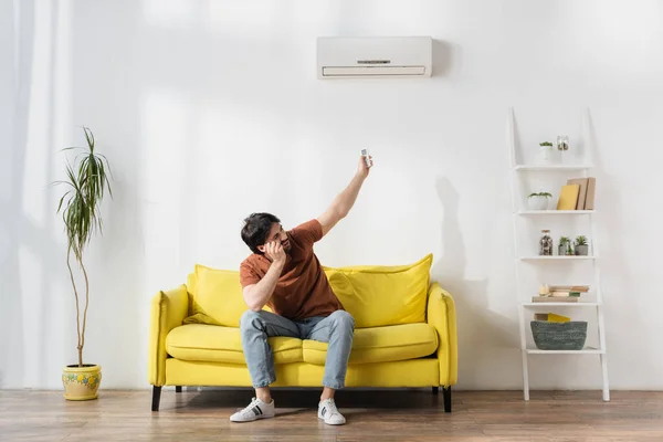 Homem usando controlador remoto perto de ar condicionado enquanto sofre de calor na sala de estar — Fotografia de Stock