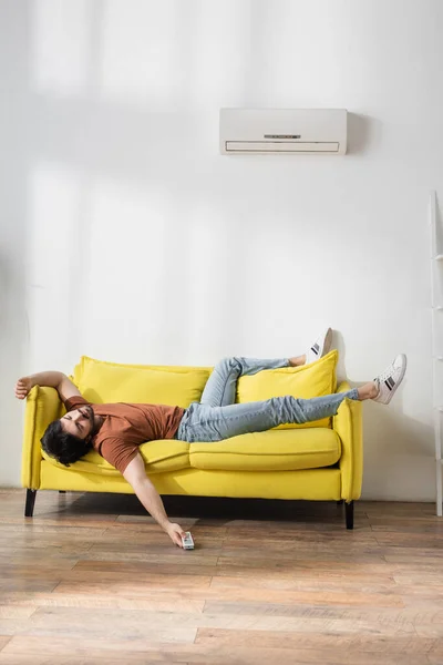Hombre sosteniendo mando a distancia mientras está acostado en el sofá y sufriendo de calor en la sala de estar - foto de stock