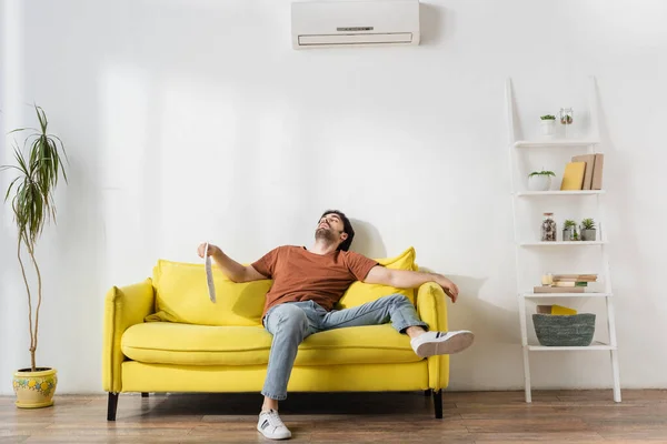 Hombre sosteniendo el periódico mientras está acostado en el sofá amarillo y sufriendo de calor en la sala de estar - foto de stock