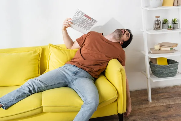 Человек машет газетой, лежа на желтом диване и страдая от жары — стоковое фото