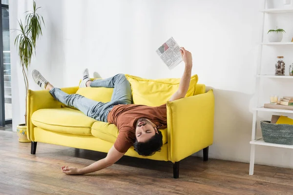 Bearded man waving with newspaper while lying on yellow couch and suffering from heat — Stock Photo