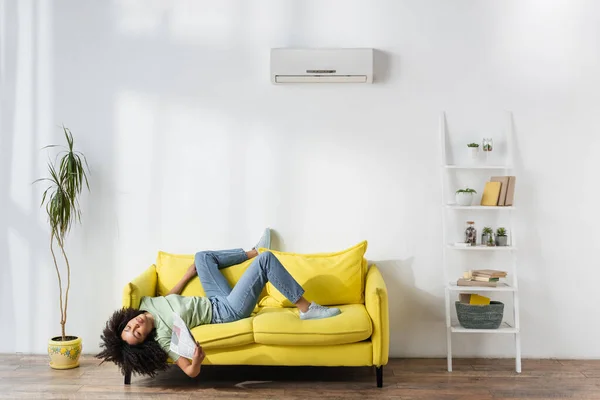 Young african american woman waving with newspaper while lying on yellow couch and suffering from heat — Stock Photo