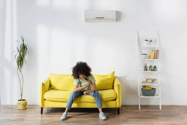 Displeased african american woman holding remote controller while sitting on yellow sofa and suffering from heat — Stock Photo