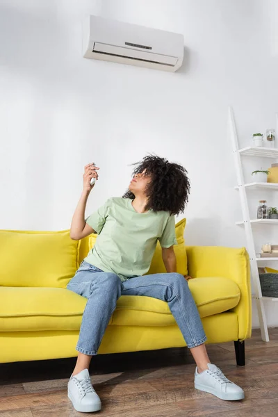 Mujer afroamericana decepcionada sosteniendo mando a distancia mientras se sienta en el sofá amarillo y sufre de calor - foto de stock