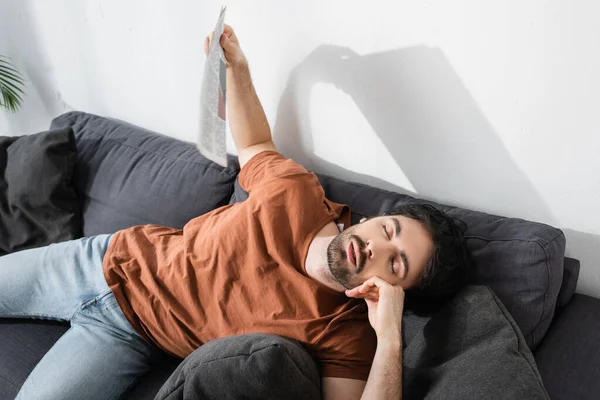 Vista de ángulo alto del hombre con los ojos cerrados ondeando con el periódico mientras está acostado en el sofá gris y sufriendo de calor - foto de stock