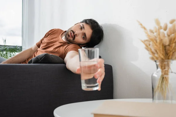 Homme barbu atteignant verre d'eau sur la table basse floue tout en souffrant de chaleur — Photo de stock