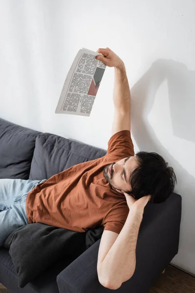 Hombre saludando con el periódico mientras está acostado en el sofá gris y sufriendo de calor - foto de stock