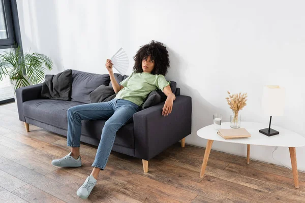 African american woman waving with hand fan while sitting on grey couch and suffering from heat — Stock Photo