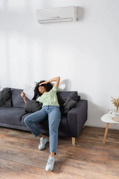 Молода афроамериканка махає ручним вентилятором, лежачи на сірому дивані і страждаючи від спеки — стокове фото