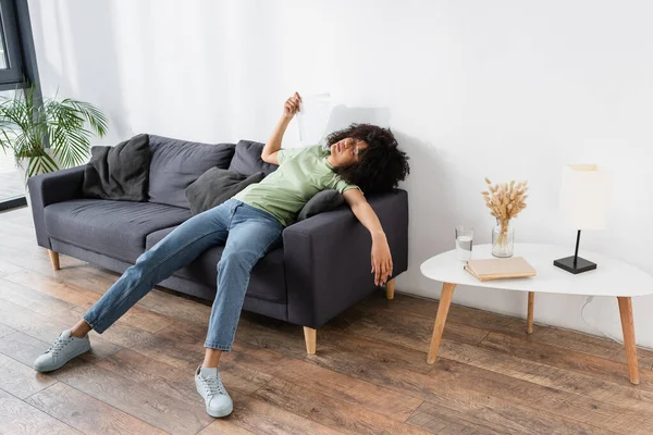 Mujer americana africana rizada agitando con ventilador de mano mientras que miente en sofá gris y que sufre de calor - foto de stock