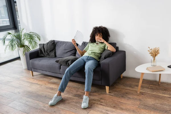 Mujer afroamericana saludando con ventilador de mano mientras bebe agua y sentada en un sofá gris - foto de stock