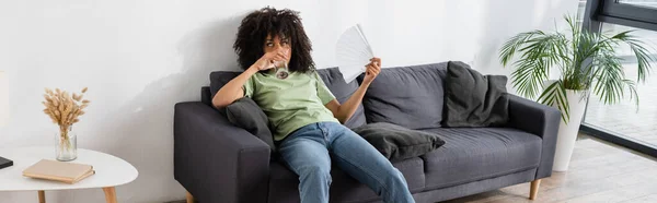 Donna afroamericana che sventola con ventilatore a mano mentre beve acqua e siede sul divano grigio, striscione — Foto stock