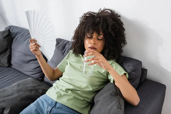 Mujer afroamericana saludando con ventilador de mano mientras sostiene un vaso de agua y se sienta en un sofá gris - foto de stock