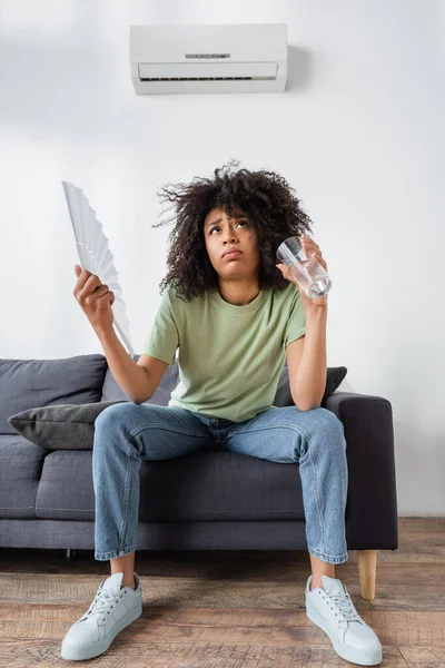 Vista de ángulo bajo de la mujer afroamericana agitando con ventilador de mano mientras sostiene el vaso de agua y sentado en el sofá gris - foto de stock