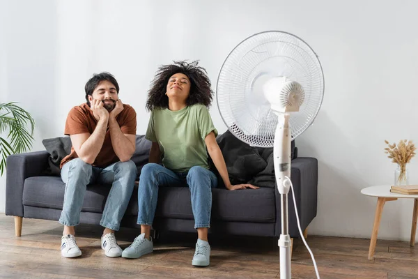 Pleased interracial man and woman sitting on couch near blurred electric fan in living room — Stock Photo