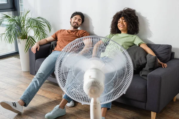 Cansado e sorrindo casal interracial sentado no sofá perto de ventilador elétrico borrado na sala de estar — Fotografia de Stock