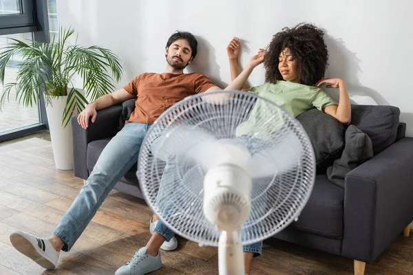 Esausto interrazziale coppia seduta su divano vicino sfocato ventilatore elettrico in soggiorno — Foto stock