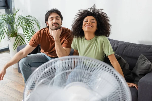 Alegre interracial pareja sentado en sofá cerca borrosa eléctrico ventilador en sala de estar - foto de stock