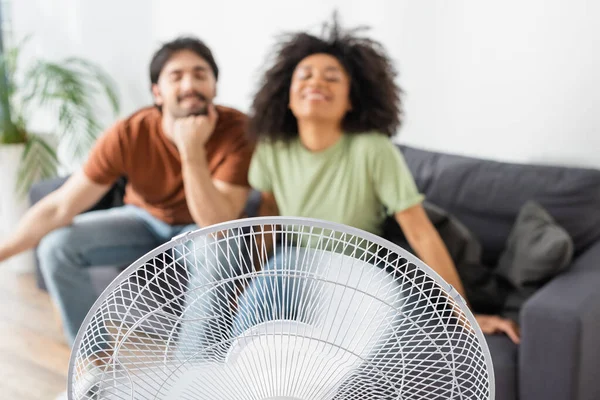 Borrosa y feliz pareja interracial sentado en el sofá cerca de ventilador eléctrico en sala de estar - foto de stock