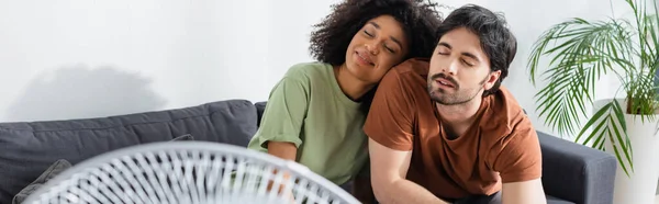 Pleased interracial couple sitting on couch near blurred electric fan in living room, banner — Stock Photo